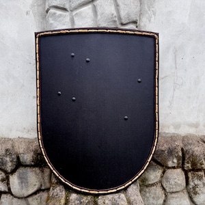 May include: A black shield with a rounded bottom and a brown border. The shield has six small, evenly spaced rivets. The border is a lighter brown color and appears to be stitched. The shield is set against a stone wall.