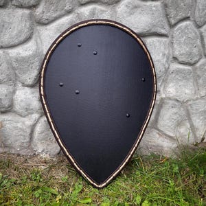 May include: A black teardrop-shaped shield with a brown and beige border. The shield has several rivets and is resting on grass in front of a stone wall. The shield is likely for historical reenactment or display.