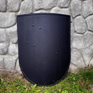 May include: A black, shield-shaped object with a slightly curved top and rounded bottom. The shield has several rivets and is set against a stone wall and grass.
