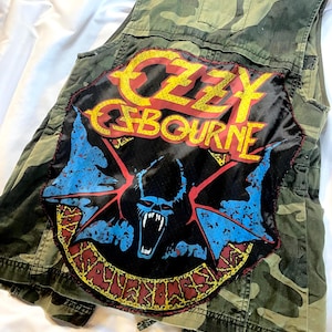 May include: Camouflage vest featuring a large patch with the text "OZZY OSBOURNE" in yellow and red, and a bat graphic with blue wings. The vest has a classic military-style design.