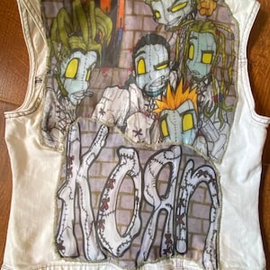 May include: White denim vest featuring a colorful graphic of cartoon figures and the band name "Korn." The artwork is stitched onto the back of the vest, with a brick wall background. The vest has a collared neckline and button closures.