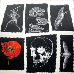 Può includere: Una collezione di quadrati di tessuto nero con illustrazioni bianche. Le illustrazioni includono uno scheletro, volpi, una lucertola, un papavero rosso, un teschio e un pipistrello. Ogni immagine ha un bordo decorativo a zig-zag.
