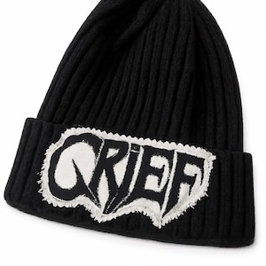 Pode incluir: Gorro preto de malha canelada com um patch branco que diz "GRIEF" em uma fonte estilizada. O patch tem uma borda irregular e desgastada. Um acessório de inverno.