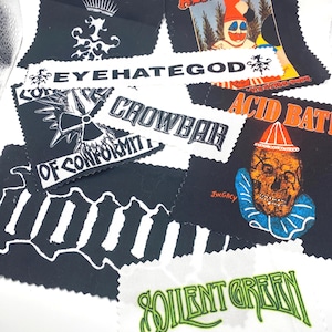 Puede incluir: Una colección de parches de tela en blanco y negro con logotipos y obras de arte de bandas. Los parches incluyen los nombres de las bandas EYEHATEGOD, CROWBAR y ACID BATH. Los parches presentan varios diseños, incluyendo una corona, una calavera y un payaso.