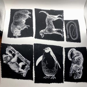May include: Six black fabric squares with white illustrations of animals. The collection includes a bear, monkey, snake, and penguin, each depicted in a detailed, vintage style. The squares are arranged on a white surface.