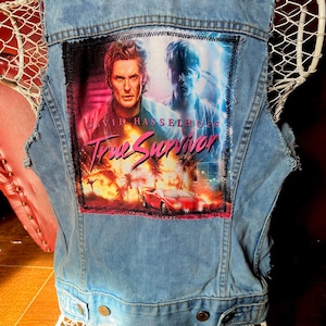 Puede incluir: Chaleco vaquero azul claro con un parche colorido que presenta a David Hasselhoff y el texto "True Survivor". El parche muestra un coche rojo, palmeras y un paisaje urbano. El chaleco tiene un aspecto desgastado con bordes deshilachados y botones metálicos.