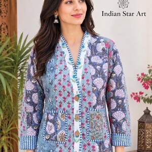 Puede incluir: Una chaqueta acolchada con base azul y estampado floral en tonos rosa, blanco y azul oscuro. La chaqueta tiene cuello en V, tres botones de madera, dos bolsillos delanteros y ribetes a rayas azules y blancas. El texto dice: Made by Indian Star Art.