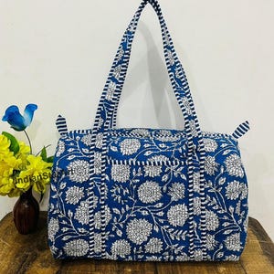 May include: A blue tote bag with a white floral pattern. The bag has two handles and a rectangular shape. The floral design covers the entire bag, including the handles. The bag is made of a quilted fabric.