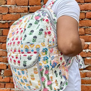 Könnte beinhalten: Ein weißer gesteppter Rucksack mit Blumenmuster. Der Rucksack hat einen schwarz-weißen Streifenbesatz und eine Reißverschlusstasche auf der Vorderseite.