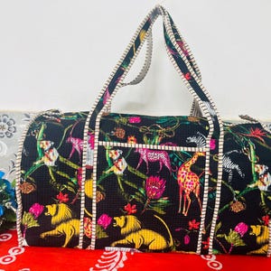 Op de afbeelding: Zwarte duffeltas met een levendige safari-dierenprint met leeuwen, giraffen en zebra's. De tas heeft crèmekleurige randen en handvatten en een ritssluiting. De tas is ongeveer 51 cm lang.