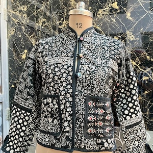 May include: Black quilted jacket with a white floral print. The jacket has a mandarin collar and a button closure. The jacket is made of a patchwork of different fabrics.