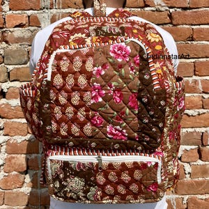 May include: A brown quilted backpack with floral and paisley patterns. The backpack has a zippered pocket on the front and two side pockets. The backpack is made of cotton and is lined with a brown fabric.