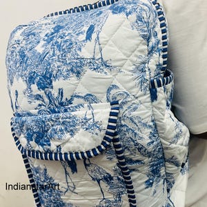 May include: White and blue quilted backpack with a toile pattern featuring birds and foliage. The backpack has a flap pocket and is trimmed with blue and white striped piping. The straps and handle are white.