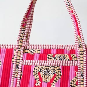 May include: A quilted tote bag with a vibrant pink and red striped pattern. The bag features tiger face designs and striped handles. The handles and trim have a white and red striped pattern.