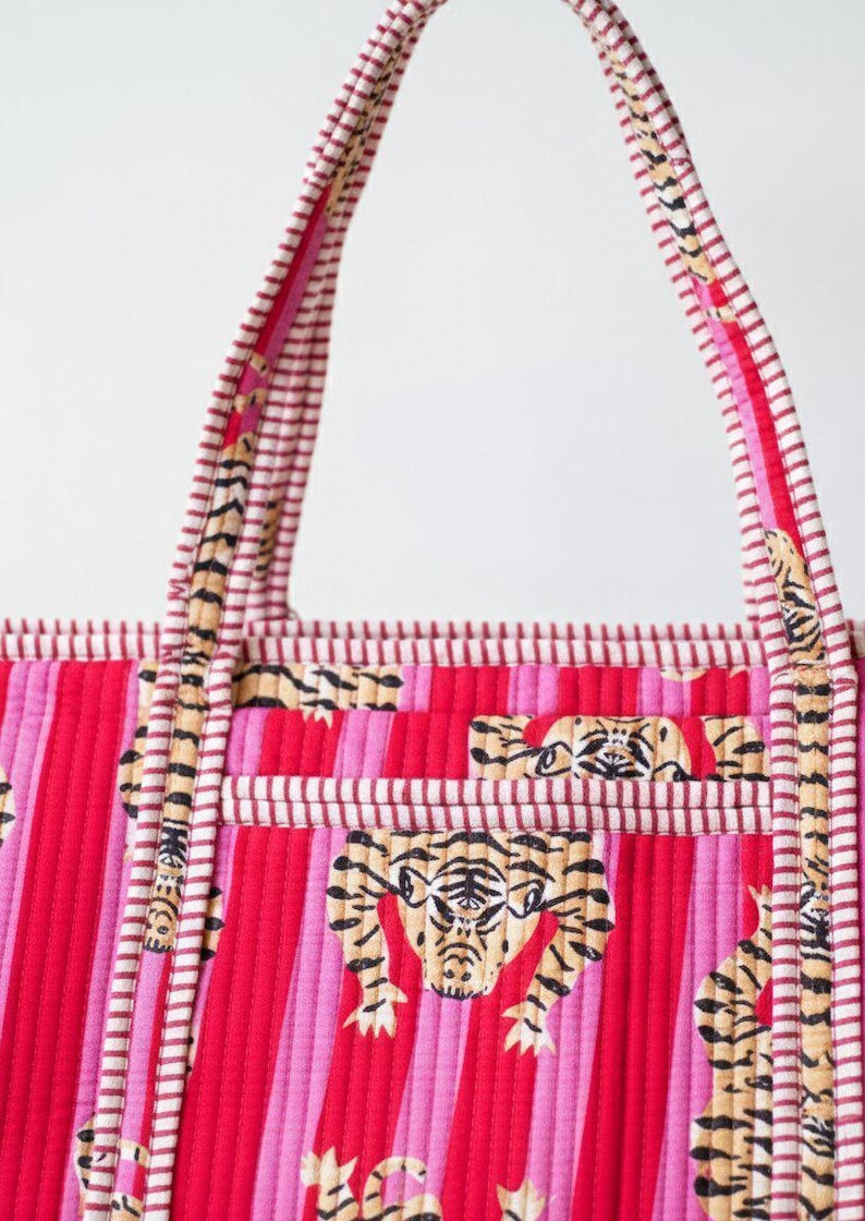 May include: A quilted tote bag with a vibrant pink and red striped pattern. The bag features tiger face designs and striped handles. The handles and trim have a white and red striped pattern.
