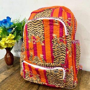 May include: Quilted backpack with a vibrant tiger stripe pattern in orange, pink, and black. Features white zippers and a side pocket. The backpack has a padded design and is suitable for carrying books and other items.