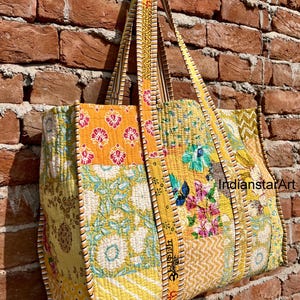 Puede incluir: Una gran bolsa de mano amarilla de patchwork con motivos florales y geométricos. La bolsa tiene asas largas amarillas con rayas blancas y negras. La bolsa está hecha de tela acolchada y tiene un aspecto único y hecho a mano.