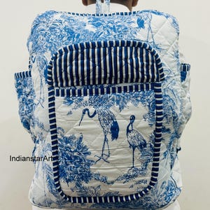 Puede incluir: Mochila acolchada blanca y azul con un diseño inspirado en la naturaleza. La mochila presenta un estampado toile de Jouy azul y blanco con pájaros y follaje. El bolsillo delantero tiene rayas verticales azules y blancas. Tiene dos bolsillos laterales y un asa superior.