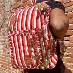 May include: A red and white striped backpack with floral embroidery. The backpack has a large pocket on the front and a drawstring closure.