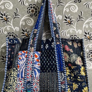 May include: A large, quilted tote bag with a patchwork design. The bag features various fabric patterns in shades of blue, red, and white. The handles and edges are trimmed with a blue and white striped fabric. The bag is hanging against a gray floral patterned wall.