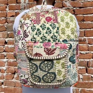 Peut inclure: Un sac à dos de style patchwork avec une grande poche avant. Le sac à dos est fait de différents tissus avec des motifs floraux et géométriques dans des tons de rose, vert et blanc. Le sac à dos a un fond crème.