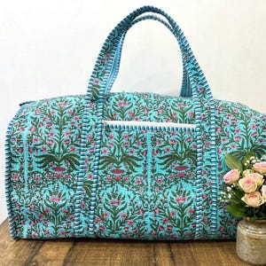 Puede incluir: Una bolsa de viaje turquesa con un estampado floral en rosa y verde. La bolsa tiene dos asas y un pequeño bolsillo. La bolsa está sobre una superficie de madera.