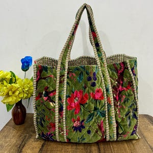 May include: A large, quilted tote bag with a vibrant floral and bird pattern in shades of green, pink, and red. The bag has sturdy handles with a striped trim and a patterned interior. The bag is sitting on a wooden surface.