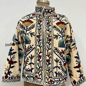 Peut inclure: Veste crème avec broderies florales complexes rouges, bleues et vertes. La veste a un col montant et une fermeture éclair. Les broderies sont détaillées avec des contours noirs, créant un contraste saisissant. La veste est présentée sur un mannequin.