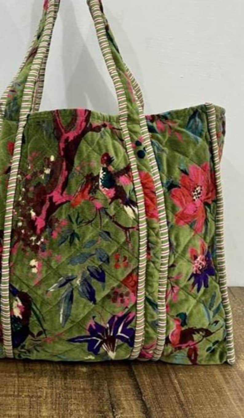 May include: Green quilted tote bag with a floral and bird pattern in shades of pink, red, and purple. The bag has striped handles and trim. The bag is made of a soft, velvety material.