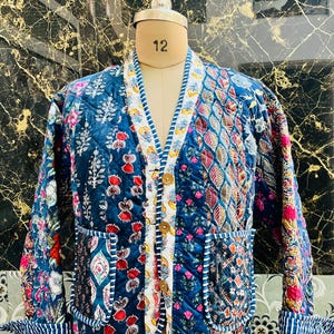 May include: A blue quilted jacket with a patchwork design featuring floral and geometric patterns. The jacket has a button closure and two pockets.