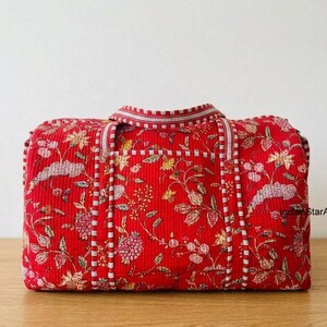 Peut inclure: Un sac de voyage matelassé rouge avec un motif floral. Le sac présente un motif répété de fleurs et de feuilles dans les tons de rose, jaune et vert. Des accents rayés blancs et rouges ornent les poignées et les bords.