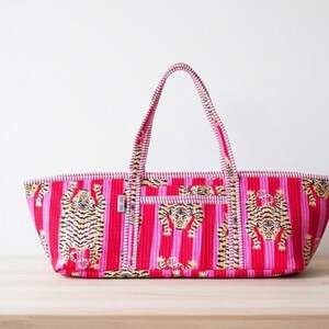 May include: A red and pink striped tote bag with tiger illustrations. The bag has a rectangular shape with a matching handle. The tiger illustrations are in black and gold. The bag is sitting on a wooden table.