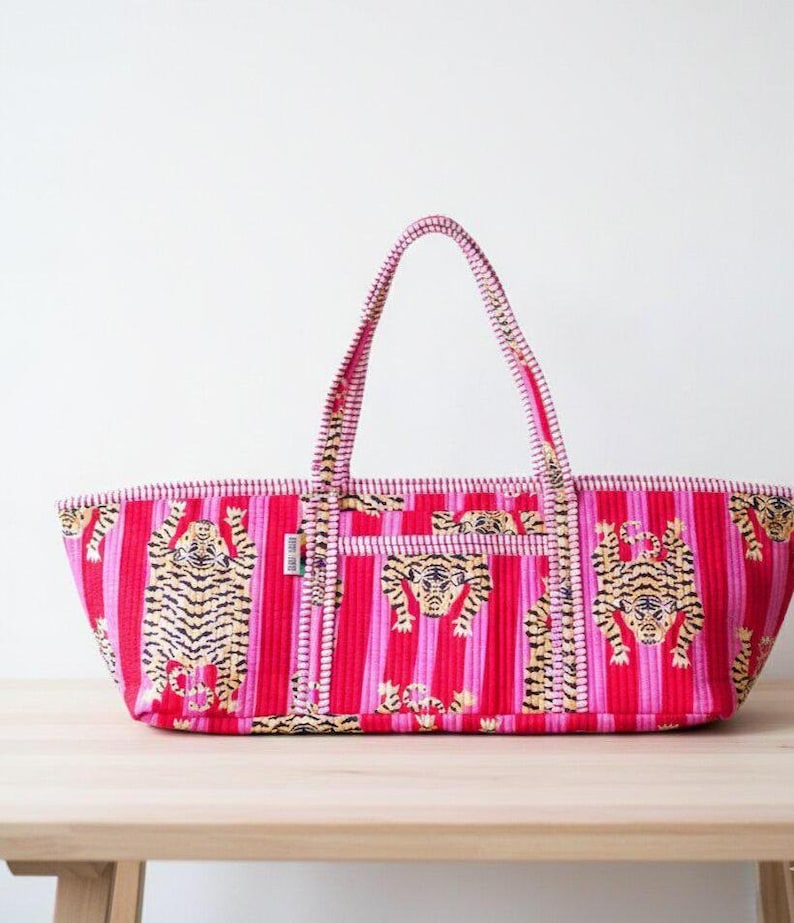 May include: A red and pink striped tote bag with tiger illustrations. The bag has a rectangular shape with a matching handle. The tiger illustrations are in black and gold. The bag is sitting on a wooden table.