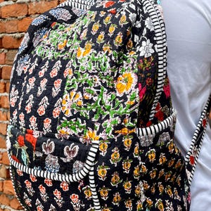 May include: A black quilted backpack with a floral print. The backpack has a white and black striped trim around the edges. The backpack is being worn by a person.