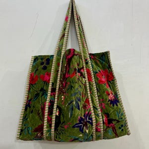 May include: Green velvet tote bag with a floral pattern featuring red, pink, and purple flowers. The bag has striped trim and handles, and a quilted design. The bag is hanging against a white wall.