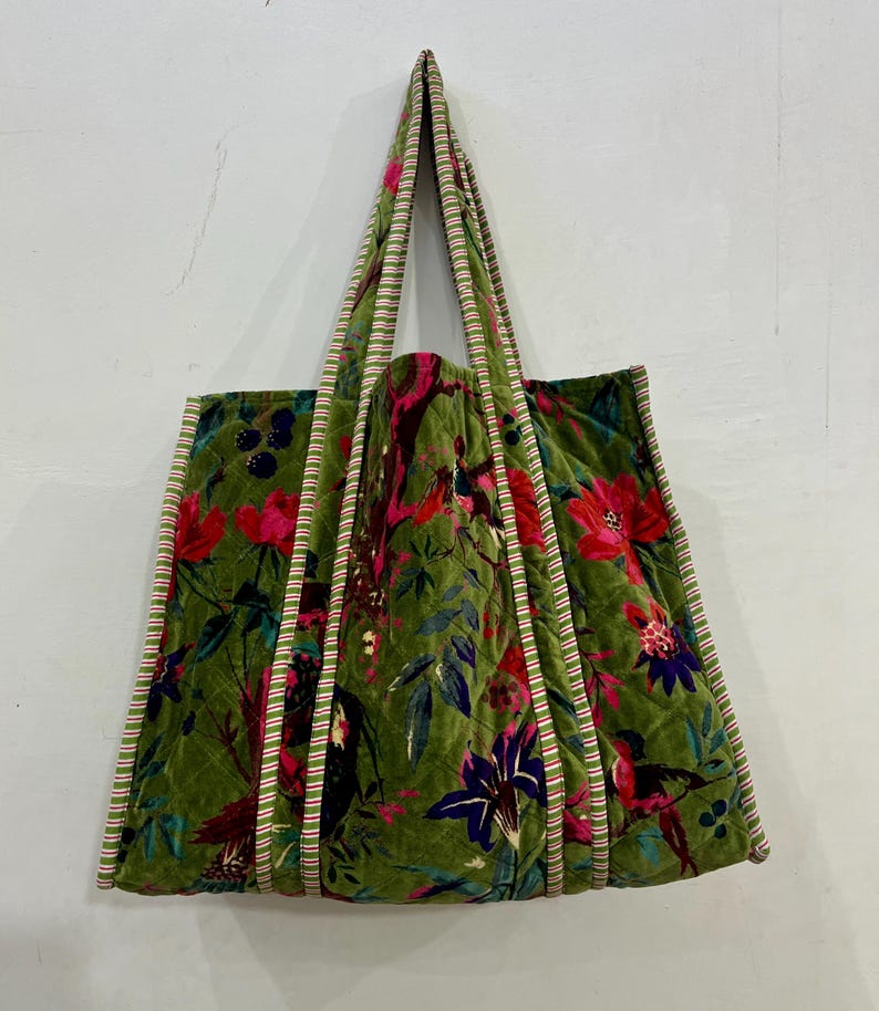May include: Green velvet tote bag with a floral pattern featuring red, pink, and purple flowers. The bag has striped trim and handles, and a quilted design. The bag is hanging against a white wall.