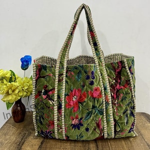 May include: A large, quilted tote bag with a floral pattern in shades of green, red, and purple. The bag has two handles and a striped trim. The bag is sitting on a wooden surface.