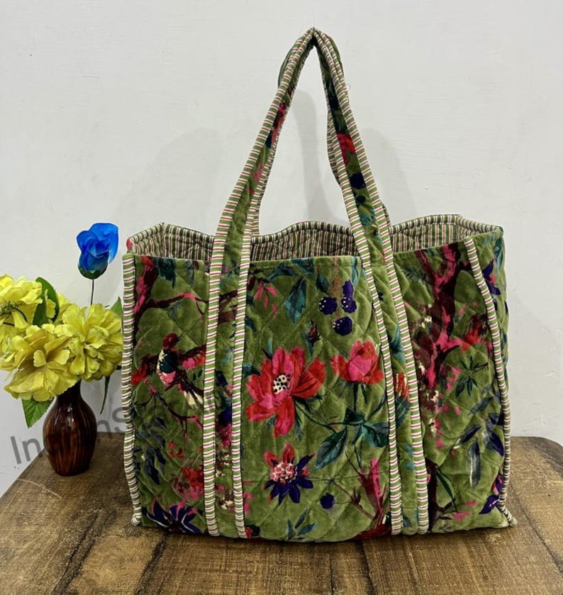 May include: A large, quilted tote bag with a floral pattern in shades of green, red, and purple. The bag has two handles and a striped trim. The bag is sitting on a wooden surface.