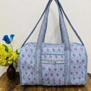 May include: A quilted duffel bag featuring a light blue base with a repeating pink and blue floral block print pattern. The bag includes two long striped shoulder straps and a front exterior pocket with a horizontal zipper closure.