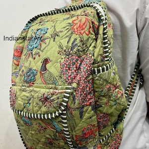 May include: Quilted backpack featuring a floral and bird design in green, pink, and blue hues. It has a white zip and black and white striped trim. The brand name "IndianstarArt" is visible on the side.