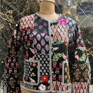 Op de afbeelding: Een gewatteerde jas met een patchwork ontwerp met bloemen en geometrische patronen in verschillende kleuren. De jas heeft een zwarte basis met wit gestreepte bies en twee voorzakken. De jas wordt op een mannequin getoond.
