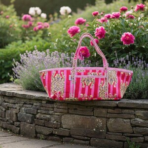 May include: A pink and red striped tote bag with tiger illustrations. The bag has a rectangular shape with a woven handle. The bag is sitting on a stone wall in front of a garden with pink flowers.