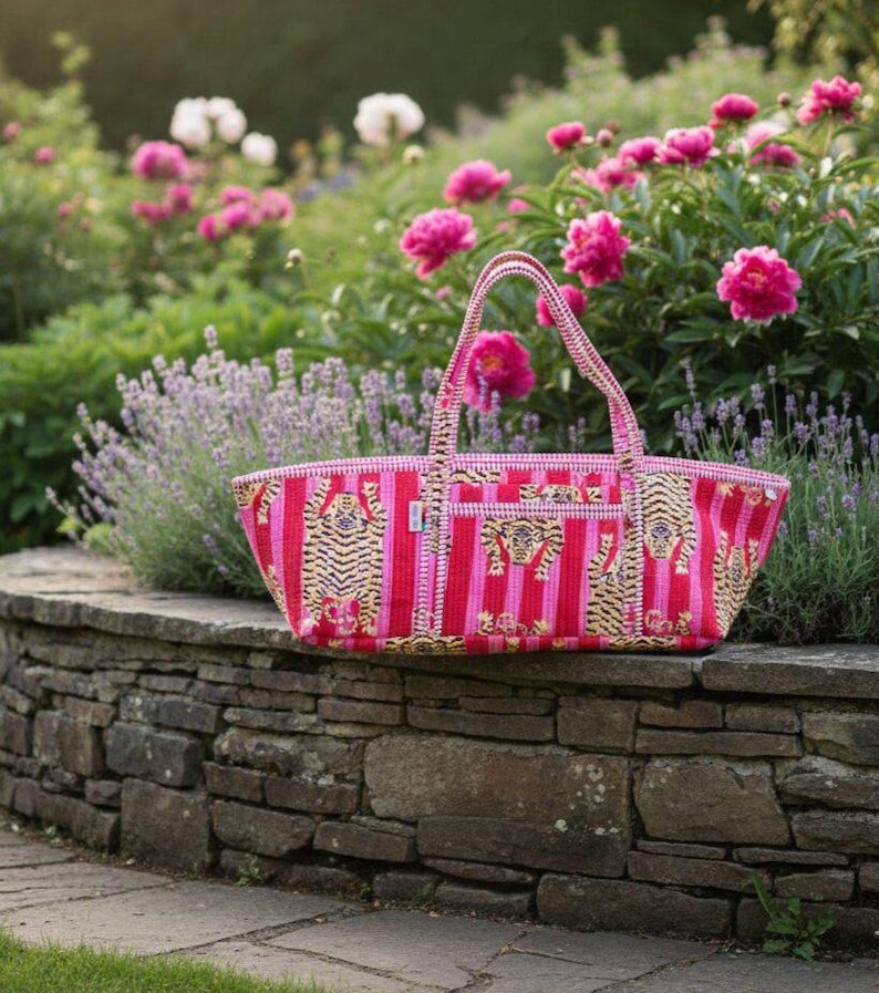 May include: A pink and red striped tote bag with tiger illustrations. The bag has a rectangular shape with a woven handle. The bag is sitting on a stone wall in front of a garden with pink flowers.