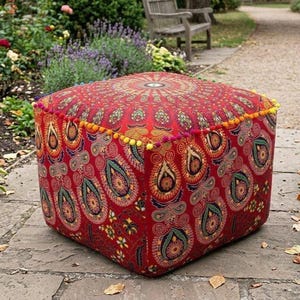 Könnte beinhalten: Ein quadratischer, roter Ottoman mit einem detaillierten Paisley-Muster in Gelb-, Grün- und Blautönen. Die Kanten sind mit bunten Pompons verziert. Dieser dekorative Hocker verleiht jedem Wohnraum eine lebendige Note.