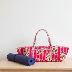May include: A red and pink striped tote bag with tiger faces and a rolled-up navy blue yoga mat. The tote bag has two handles and a rectangular shape. The yoga mat is on a wooden surface.