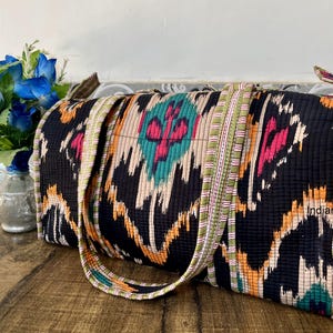 May include: A black and white ikat fabric tote bag with colorful accents. The bag has a long strap and is made of a soft, quilted fabric.