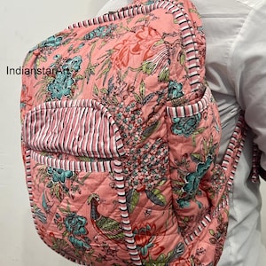 May include: A quilted backpack with a vibrant floral pattern in shades of pink, teal, and green. The backpack features a front pocket with vertical pink and white stripes and black and white striped trim. The design includes a bird and floral motifs.