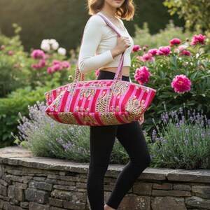 May include: A pink and red striped tote bag with a decorative pattern. The bag has two handles and is being carried outdoors. The background includes a stone wall, flowers, and greenery.