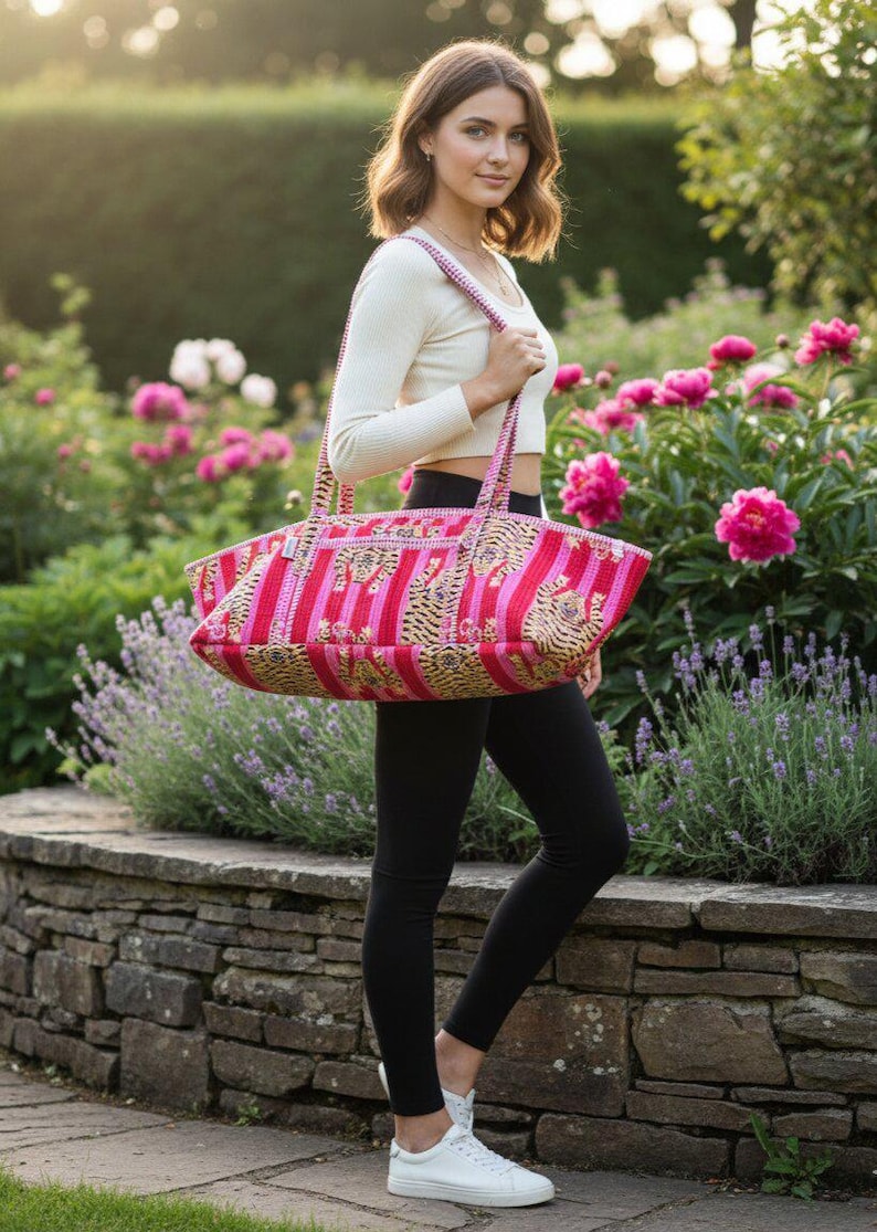 May include: A pink and red striped tote bag with a decorative pattern. The bag has two handles and is being carried outdoors. The background includes a stone wall, flowers, and greenery.