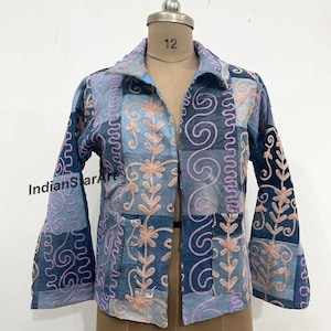 May include: A patchwork denim jacket with a collared neckline and long sleeves. The jacket features embroidered floral and swirl designs in pink thread. The jacket is displayed on a mannequin.